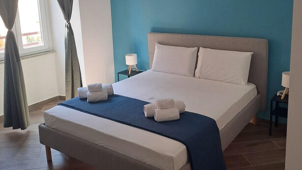 A bedroom with a bed, a bedside table, a lamp, and a window with curtains. - At Home Gaeta Guest House (Rome)