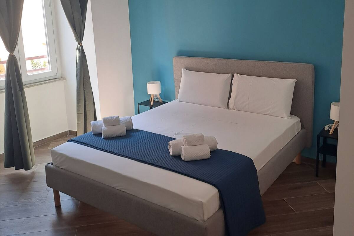 Un dormitorio con cama, mesita de noche, lámpara y ventana con cortinas.