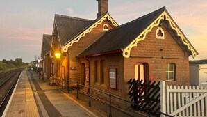 Un andén de estación de tren con un edificio de ladrillo, una cerca de piquetes blancos y una vía de tren.