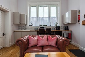 A cozy kitchen with a dining area, featuring a brown leather sofa, wooden table, and a window with curtains.
