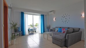 Ein Wohnzimmer mit grauem Sofa, einem Couchtisch und einer Zimmerpflanze. Ein Fenster mit blauen Vorhängen bietet einen Blick ins Freie.