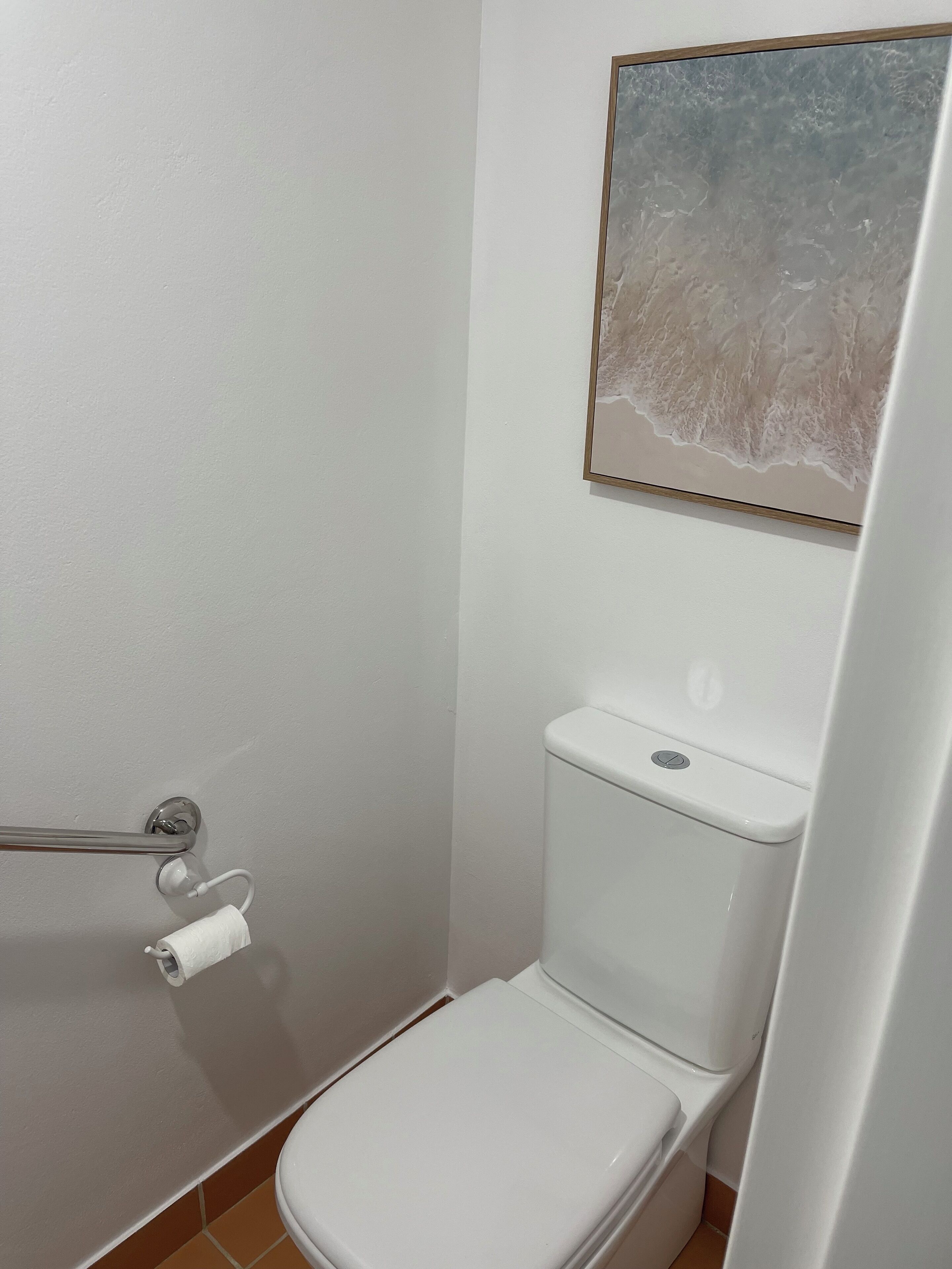 Un baño pequeño y limpio con inodoro blanco, dispensador de toallas y una obra de arte enmarcada en la pared.
