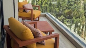 A balcony with two wooden chairs and a small table, a glass railing, and a view of buildings and trees. - Luxury golf villa with heated pool-near center (Marrakech)