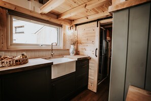 A wooden interior with a kitchen area, including a sink, a basket of bread, and a vase with dried flowers. - Rowan (Amlwch)