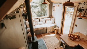 A cozy cabin interior with a wood-burning stove, a bench with cushions, a dining table with a basket of bread, and a view of the outside. - Rowan (Amlwch)