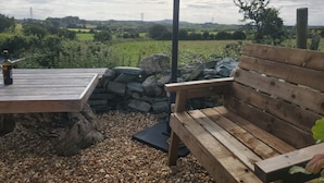 A wooden bench and table with a bottle of wine, surrounded by pebbles and stone walls, overlooking a green landscape with distant hills. - Rowan (Amlwch)