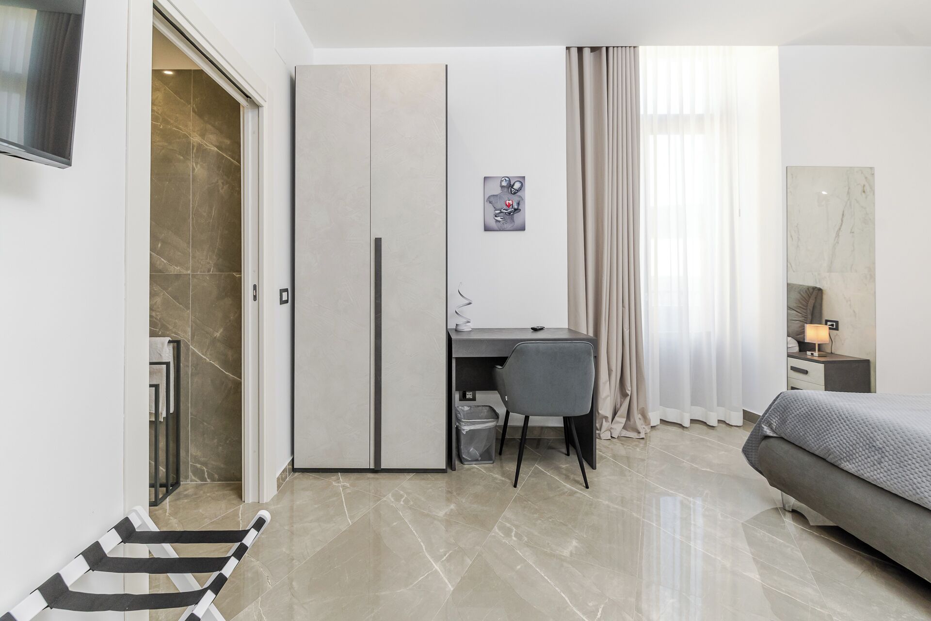 A modern bedroom with a bed, a desk with a chair, a wardrobe, and a TV mounted on the wall.