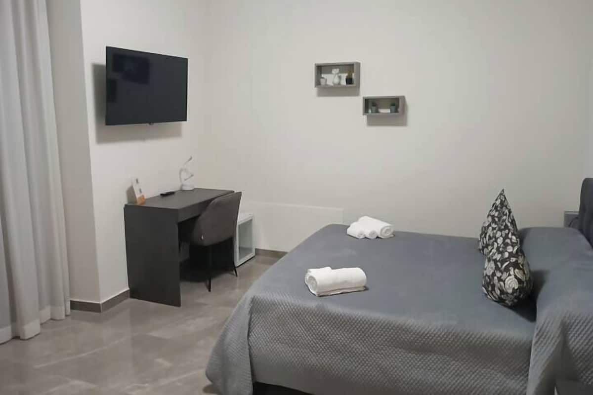 A hotel room with a bed, a desk, a TV, and a small shelf with decorative items.