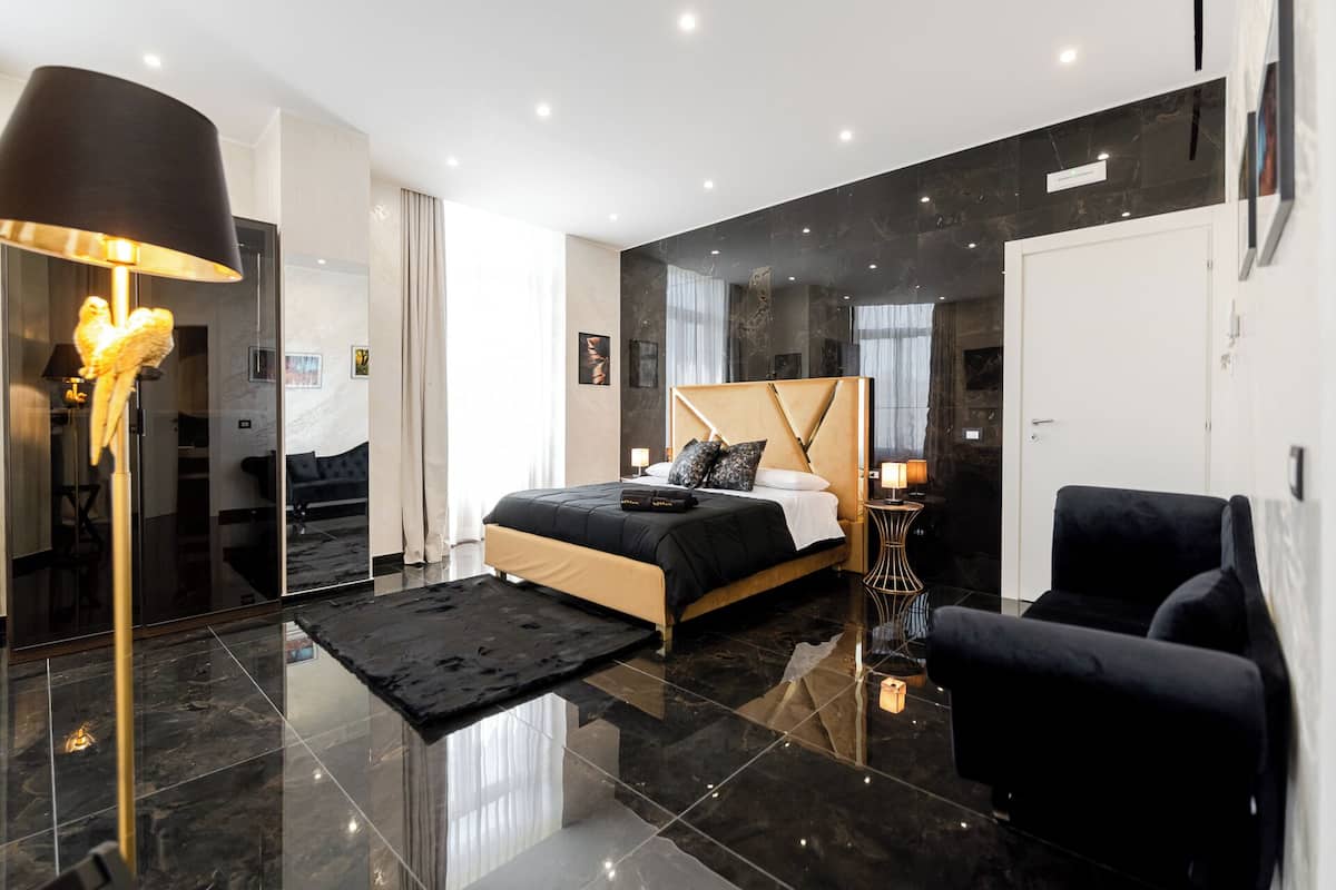 A modern bedroom with a large bed, a black armchair, a floor lamp, and a mirror.