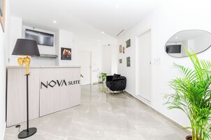 A modern reception area with a wooden reception desk labeled "NOVA SUITE," a black floor lamp, a black armchair, and a potted plant.