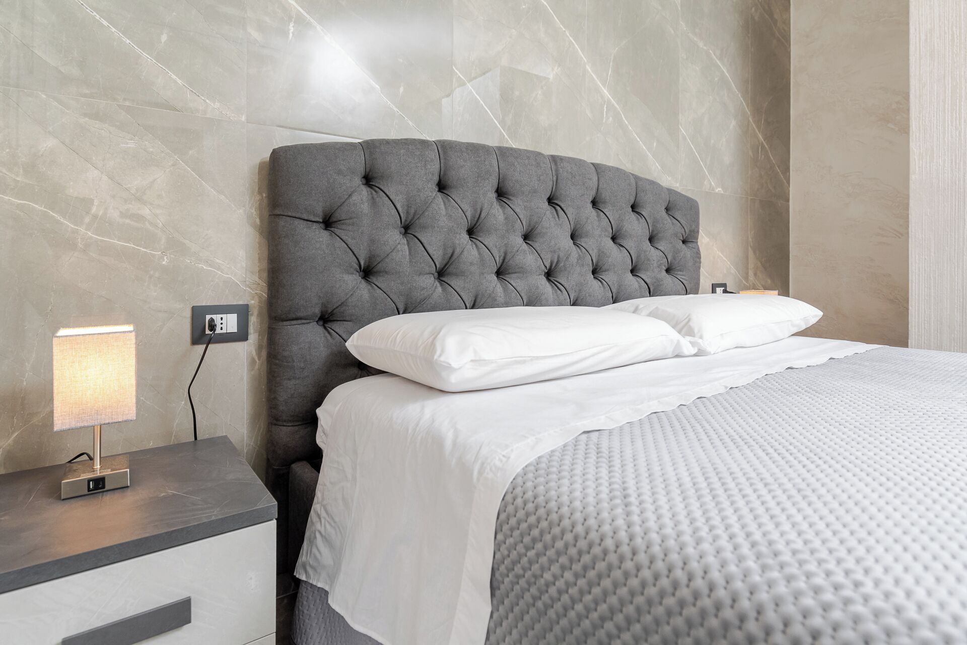 Una cama bien hecha con cabecera capitonada, ropa de cama blanca y una mesita de noche con lámpara.