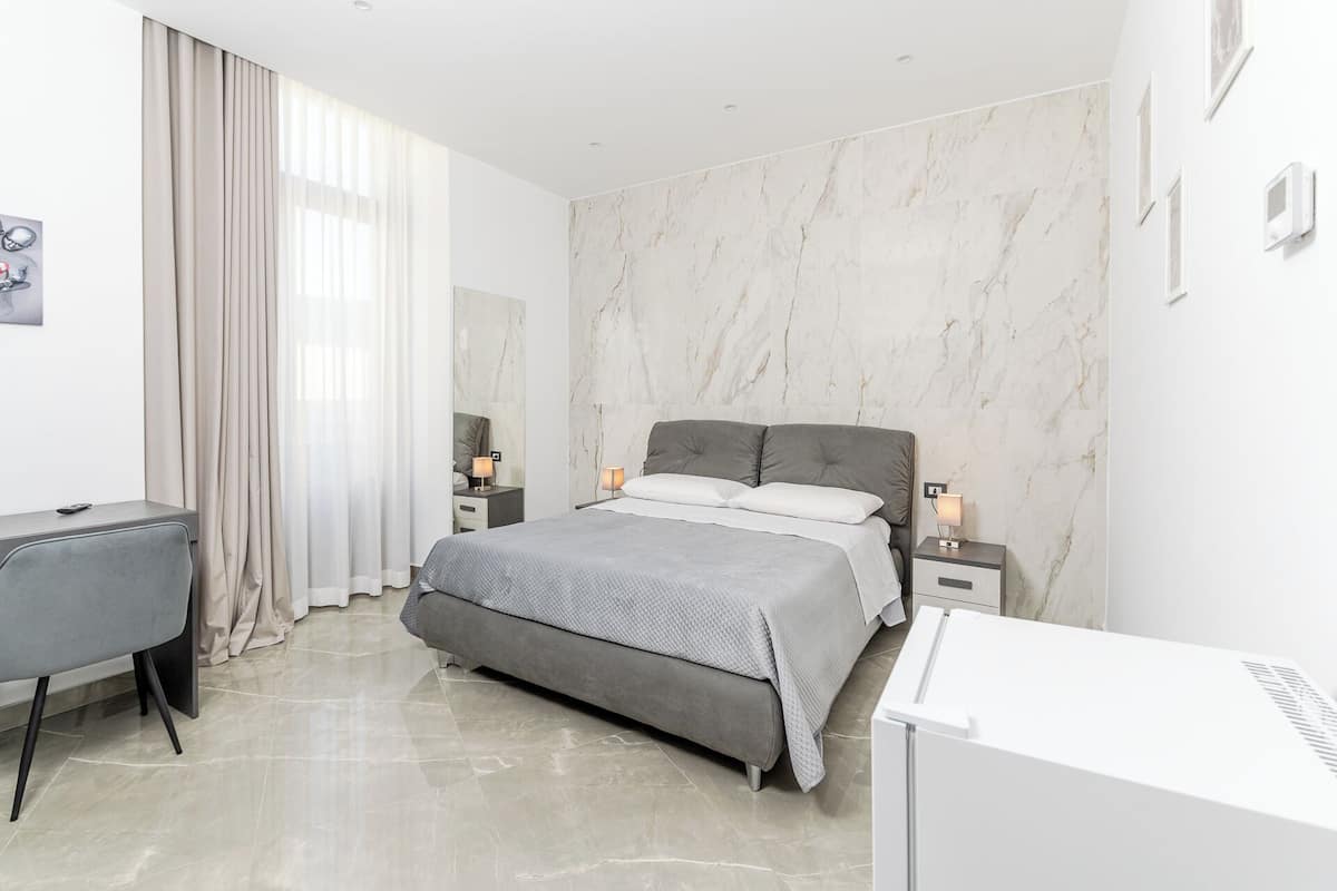 A modern hotel room with a marble wall, a grey bed, a desk, and a chair.