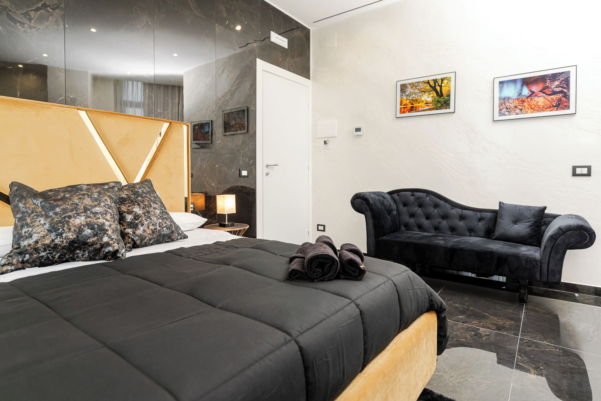A modern hotel room with a large bed, a black sofa, and two framed pictures on the wall.