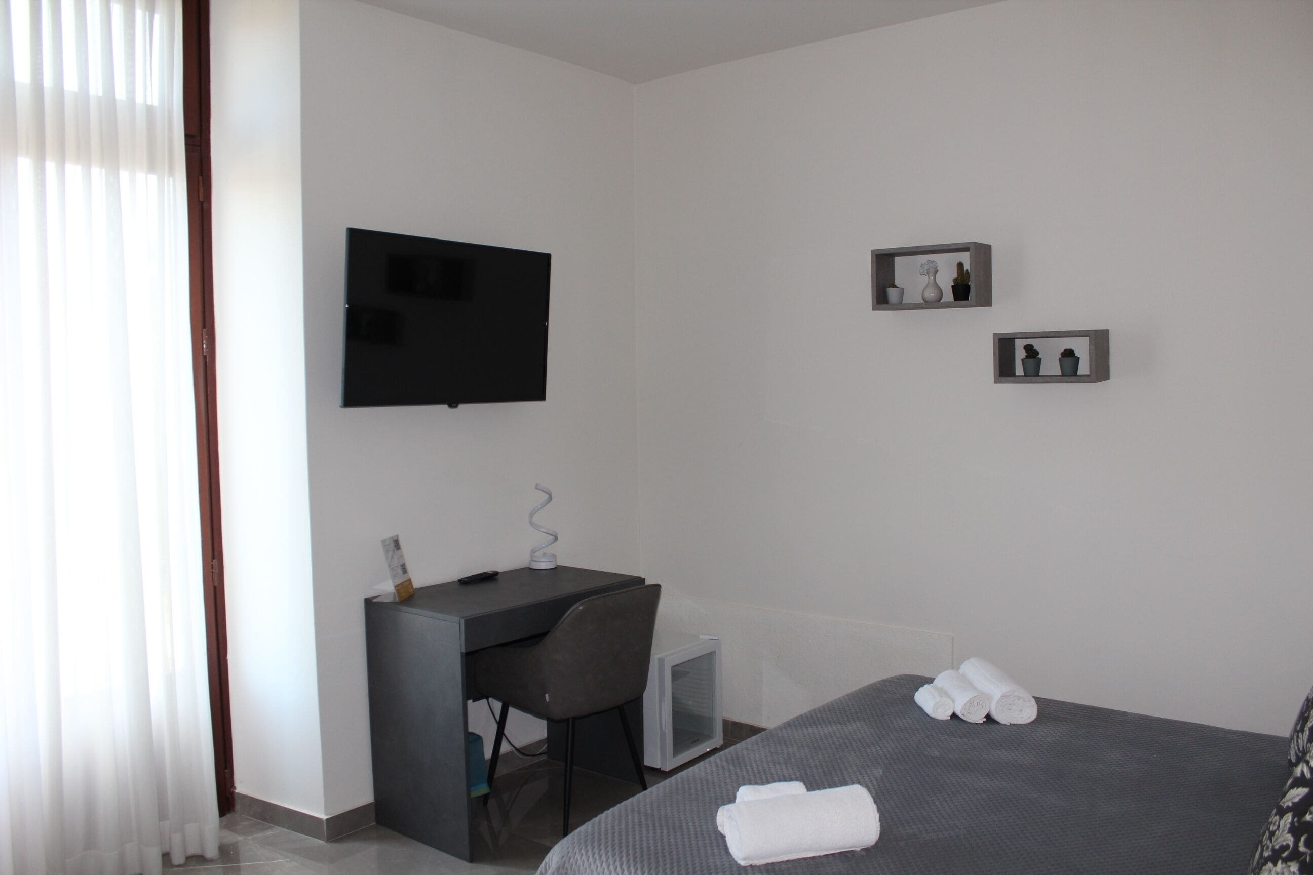 A hotel room with a bed, a desk, a chair, a television, and a small shelf with decorative items.