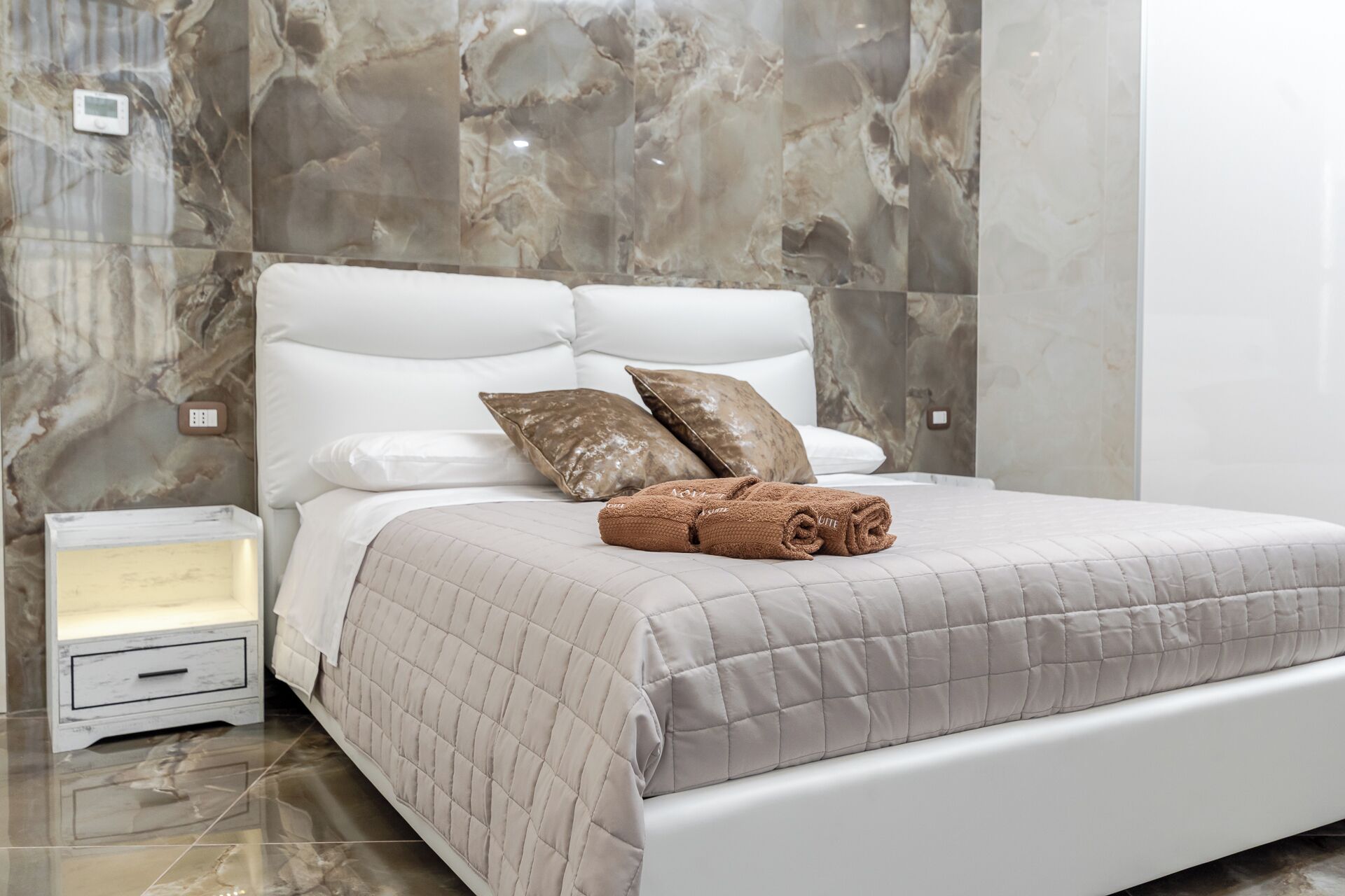 A neatly made bed with a beige quilt, two pillows, and a rolled towel on a white headboard in a room with marble walls and a small bedside table.