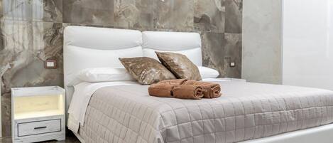 A neatly made bed with a beige quilt, two pillows, and a rolled towel on a white headboard in a room with marble walls and a small bedside table.