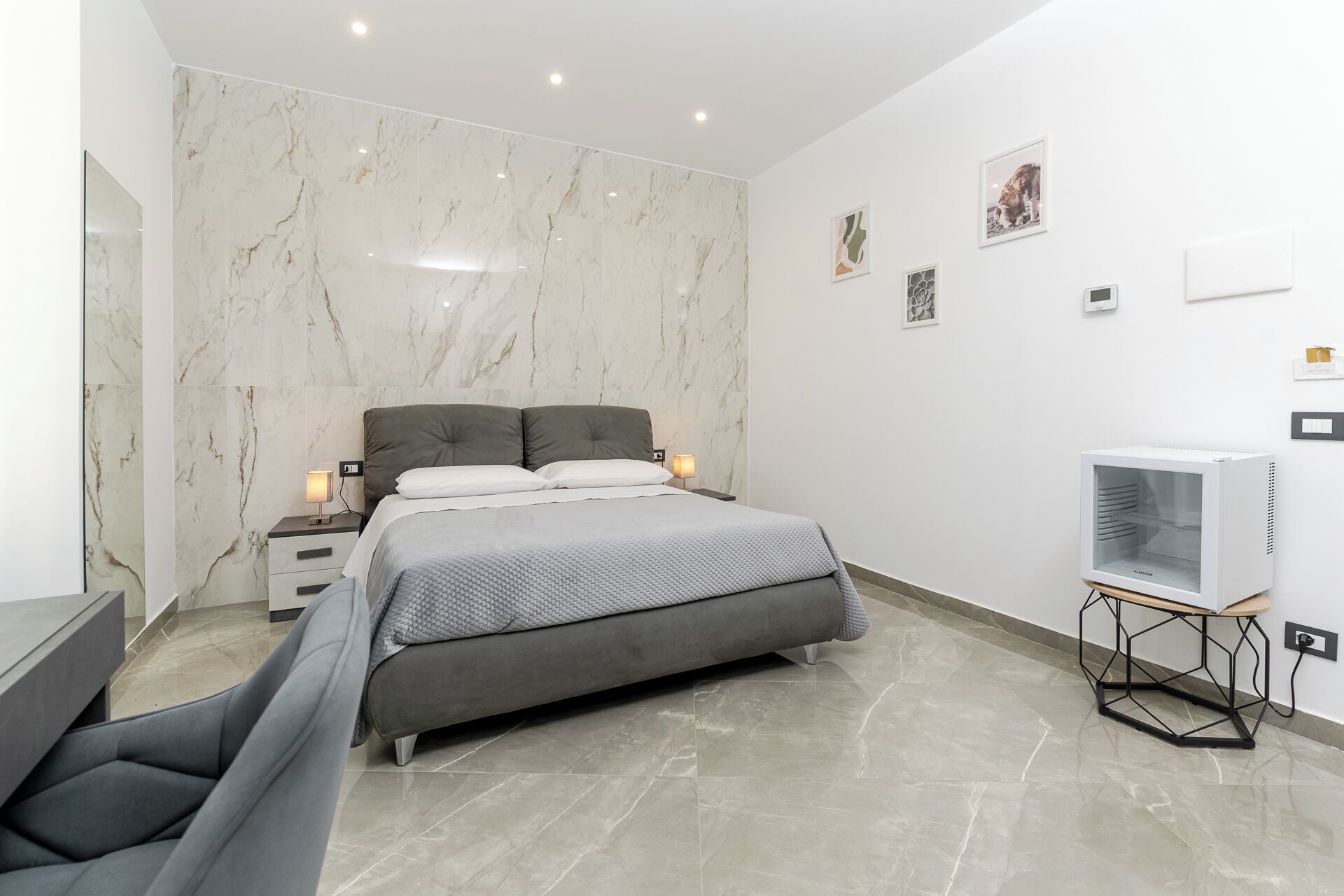 Un dormitorio moderno con pared de mármol, cama con ropa de cama gris, lámparas de noche, televisor sobre un soporte y una obra de arte en la pared.