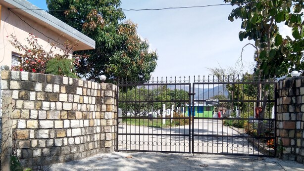 A gated entrance with a stone wall and metal gate, leading to a paved area with a building in the background.