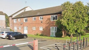 A multi-story brick building with a flat roof, a gated parking area, and a tree in front.