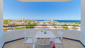 A balcony with a table and chairs, overlooking a coastal cityscape with buildings and palm trees. - Sea View Apartment Skyline by the Sea (Playa de Las Americas)