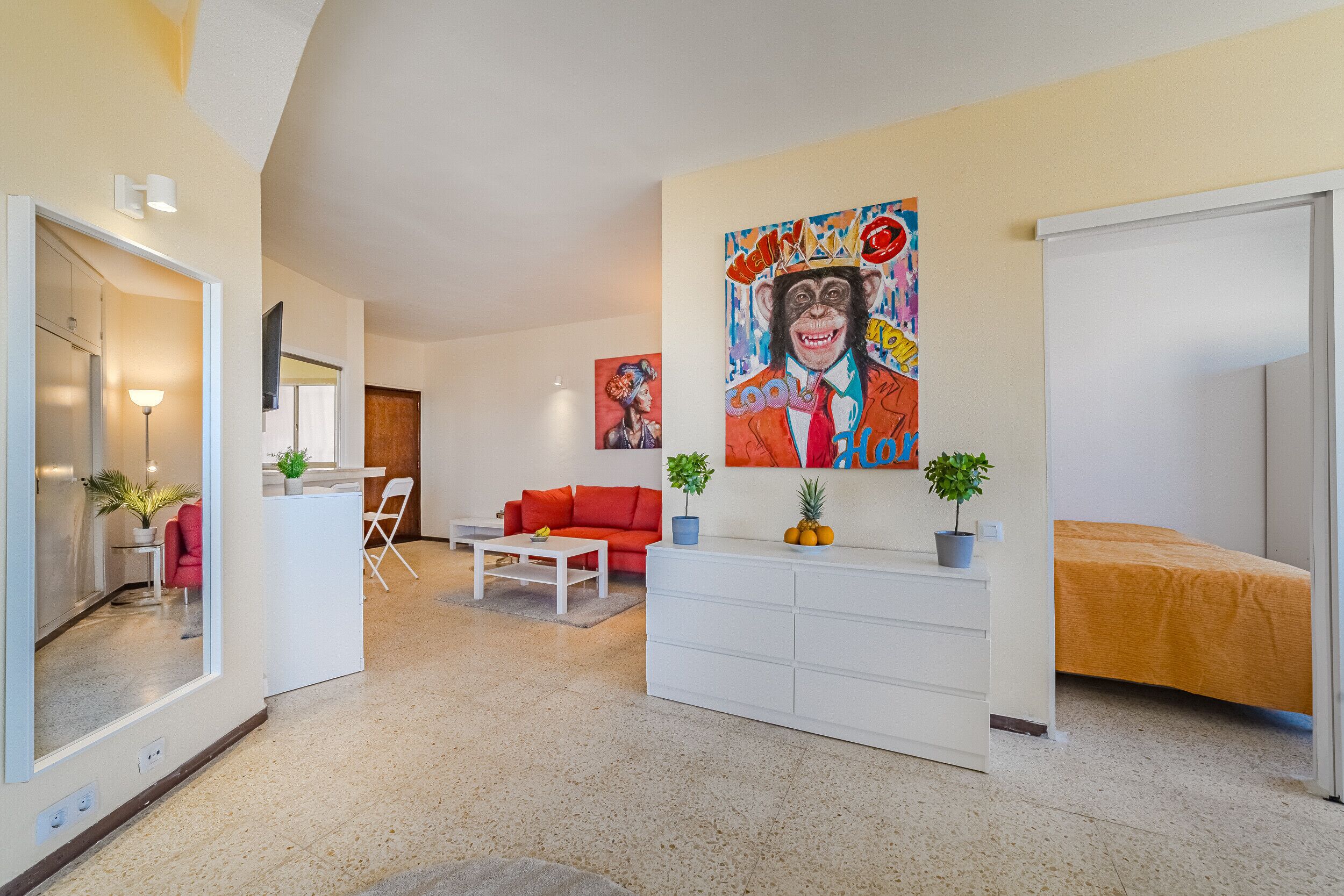 Un salon moderne avec un canapé rouge, une table basse blanche et un grand tableau représentant un singe accroché au mur. Une petite table avec une plante et un miroir se trouve sur le côté gauche.