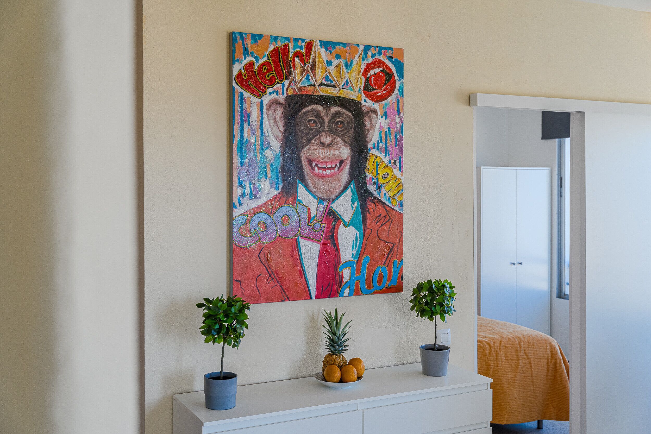 A room with a colorful painting of a monkey wearing a crown, a white dresser with two potted plants, and a bed with an orange blanket.