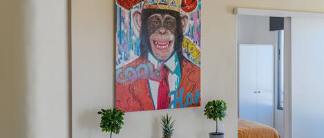 A room with a colorful painting of a monkey wearing a crown, a white dresser with two potted plants, and a bed with an orange blanket.