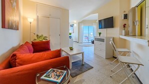 A modern living room with a red sofa, a coffee table with a magazine, a TV mounted on the wall, and a kitchen area with a dining table and chairs. - Sea View Apartment Skyline by the Sea (Playa de Las Americas)