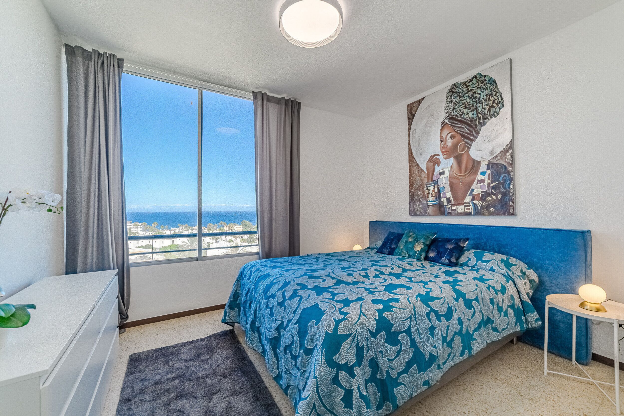 A bedroom with a large bed, a view of the city, a painting on the wall, and a bedside table with a lamp.