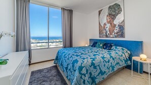 A bedroom with a large bed, a view of the city, a painting on the wall, and a bedside table with a lamp. - Sea View Apartment Skyline by the Sea (Playa de Las Americas)