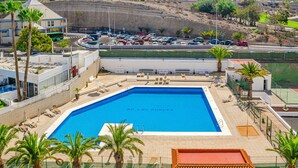 A swimming pool with a clear blue water, surrounded by palm trees and a building with a red roof in the background. - Sea View Apartment Skyline by the Sea (Playa de Las Americas)