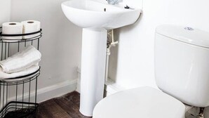 A modern bathroom with a white sink, toilet, and a mirror above the sink. There is a shelf with toiletries and a plant.