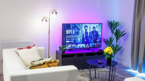 A modern living room with a white sofa, a flat-screen TV, a floor lamp, and a small round table with a plant.