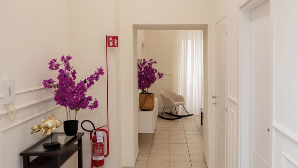 A hallway with a tiled floor, a wooden bench with a vase of purple flowers, a red fire extinguisher, and a white door at the end.