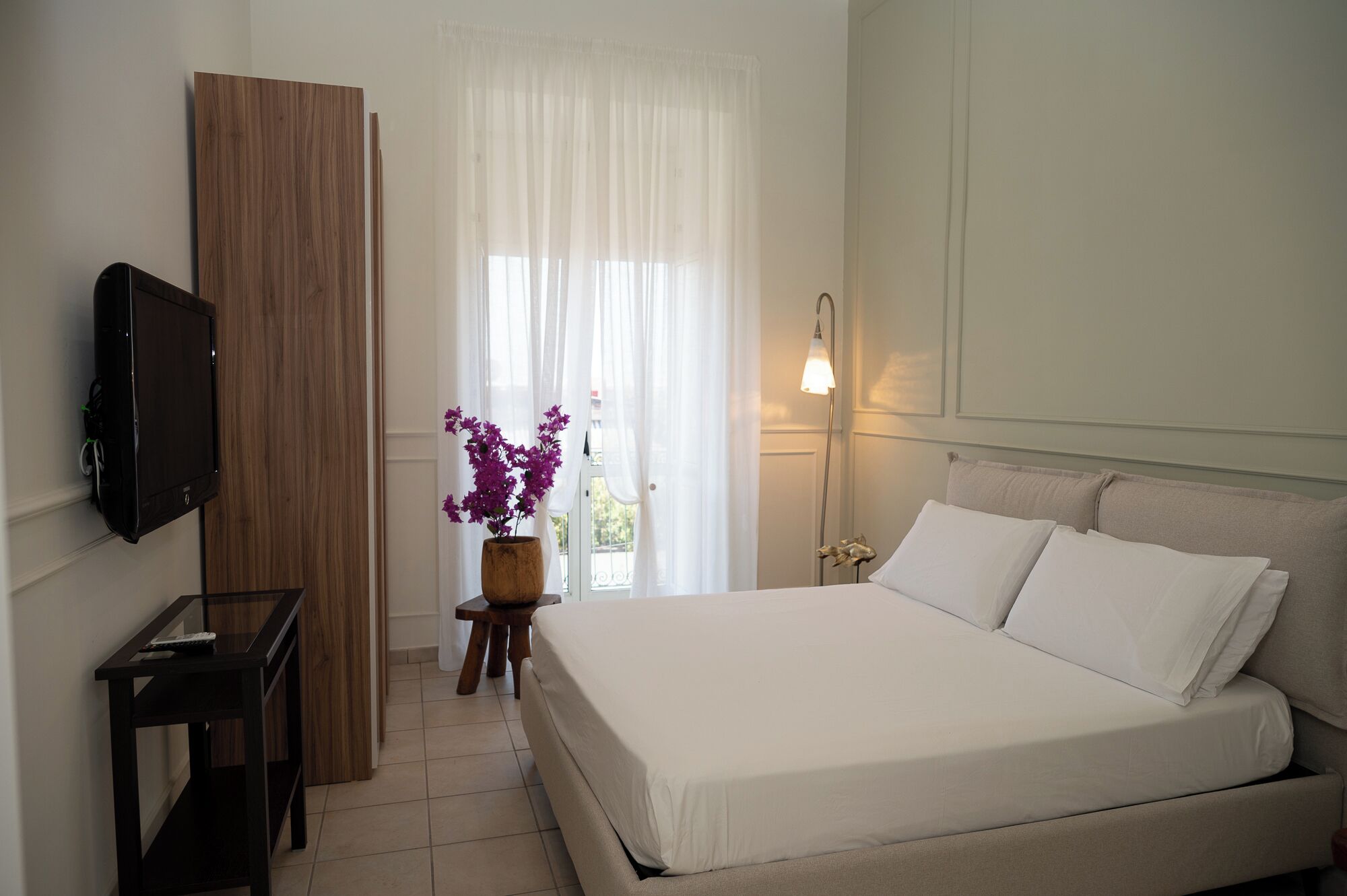 A bedroom with a bed, a TV, a wooden wardrobe, a small table with a vase of flowers, and a bedside lamp.