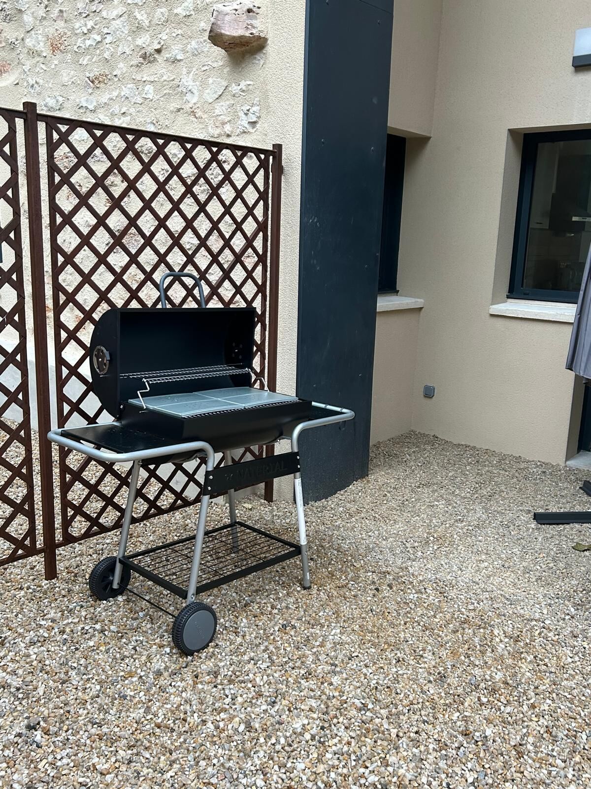 Un barbecue portatif sur roulettes, placé sur une surface de gravier, près d’un bâtiment doté d’une clôture en treillis et d’une fenêtre.