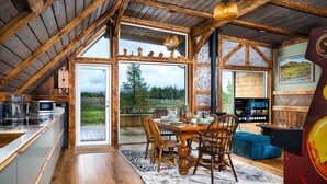 L’intérieur d’un chalet rustique comprend un coin repas en bois, une cuisine équipée d’appareils modernes et une grande fenêtre donnant sur un paysage boisé.
