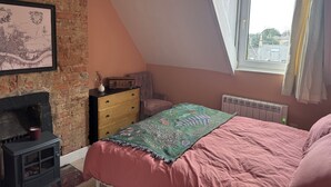 Une chambre avec un plafond en pente, une cheminée, un lit recouvert d’une couverture rose, une commode et une fenêtre avec des rideaux.