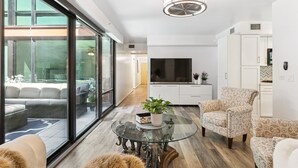 Un salon moderne avec une table basse en verre, un canapé et des fauteuils à motifs. Un téléviseur à écran plat est fixé au mur, et une plante en pot est posée sur la table.