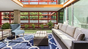 Un intérieur moderne avec un plafond haut, un coin salon comprenant un canapé et un pouf, une petite table et une vue sur un immeuble rouge doté de grandes fenêtres.
