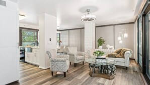 Un salon moderne avec une table basse ronde en verre, des fauteuils à motifs, un canapé blanc et une grande fenêtre donnant sur l’extérieur.