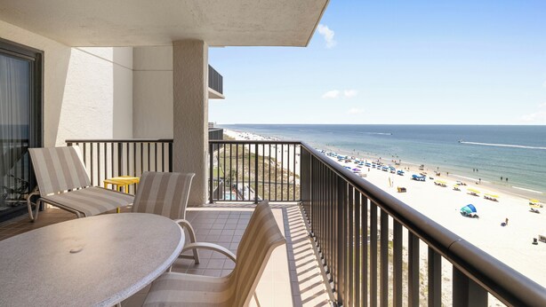 Ein Balkon mit Tisch und Stühlen mit Blick auf einen Strand mit Sonnenschirmen und Menschen.