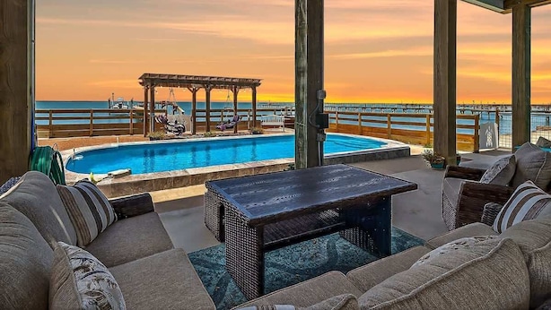 A poolside area with a pergola, lounge chairs, and a view of the ocean at sunset.