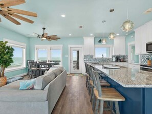 A modern kitchen with a large island, a dining area with a view, and a living room with a sofa and a plant.