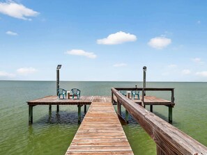 A wooden pier extending into calm water with two chairs on each end.
