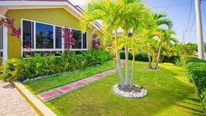 A yellow building with a well-maintained garden featuring palm trees and various plants. - 3BR Villa in Drax Hall Country Club (St. Ann's Bay)