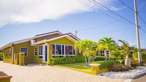 A yellow two-story house with a paved driveway, a small garden with palm trees, and a utility pole with power lines. - 3BR Villa in Drax Hall Country Club (St. Ann's Bay)