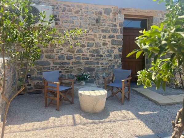 Un espace extérieur aménagé avec des murs en pierre, comprenant deux chaises en bois et une table ronde, le tout entouré de gravier et de verdure.