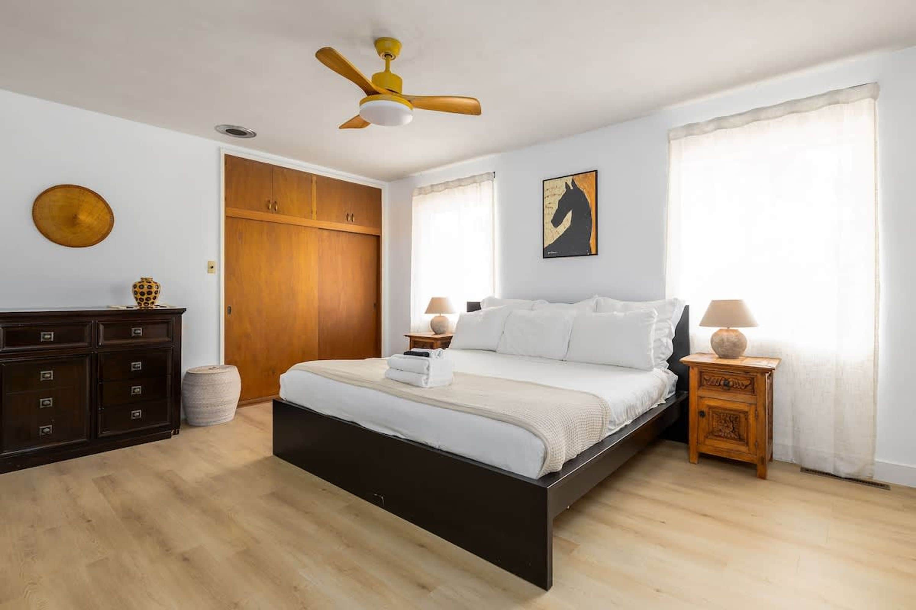 Ein Schlafzimmer mit Bett, Holzschrank, Deckenventilator und einem Fenster mit Vorhängen.