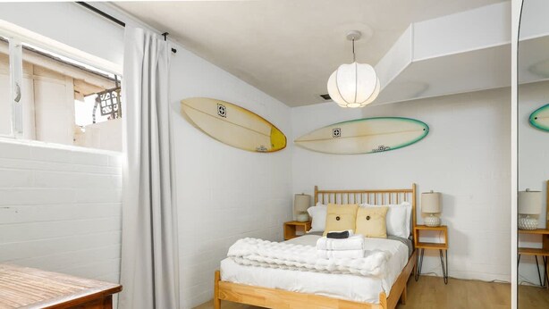 A bedroom with a wooden bed, white bedding, two surfboards on the wall, and a wooden nightstand with a lamp.